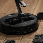 A black robot vacuum on a wooden floor surrounded by socks. A robotic arm is coming out of the center of the robot.