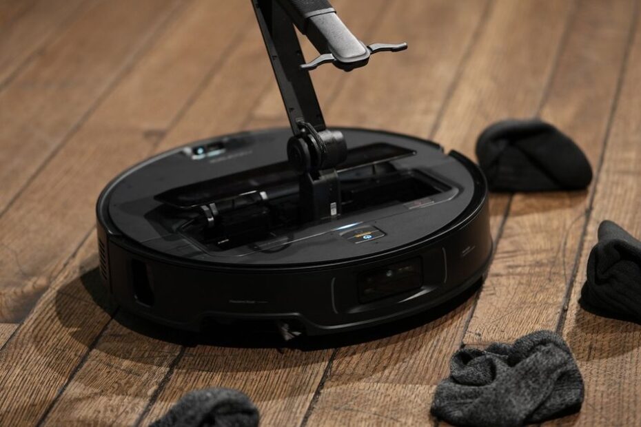 A black robot vacuum on a wooden floor surrounded by socks. A robotic arm is coming out of the center of the robot.